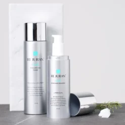 Two silver ReJURAN skincare bottles—a balancing toner and a refreshing emulsion—stood on a marble board with a rosemary sprig nearby.