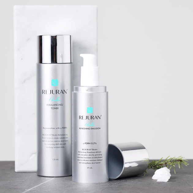 Two silver ReJURAN skincare bottles—a balancing toner and a refreshing emulsion—stood on a marble board with a rosemary sprig nearby.