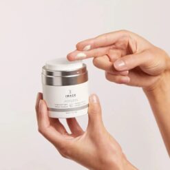 Hands holding a white jar of IMAGE Skincare Ageless Total Overnight Retinol Masque while a fingertip scoops a dab of cream from the lid edge (skincare product in use)
