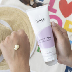 Person applying lotion from a white tube labeled Image Skincare Body Spa onto the back of the other hand, with a straw hat and colorful background nearby.