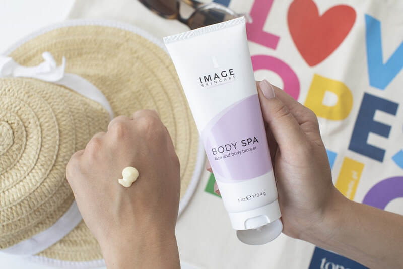 Person applying lotion from a white tube labeled Image Skincare Body Spa onto the back of the other hand, with a straw hat and colorful background nearby.