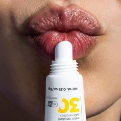 Close-up of glossy red lips with a white lip balm tube pressed against the lower lip as if applying balm.