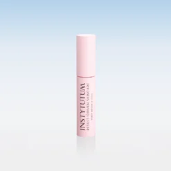 Pink cylindrical cosmetic tube standing upright on a glossy surface with a soft blue-to-white gradient background; branding reads INSTYTUTUM.