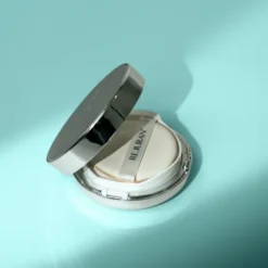 Silver round compact powder case opened to reveal white powder labeled REJURAN on a light turquoise background.