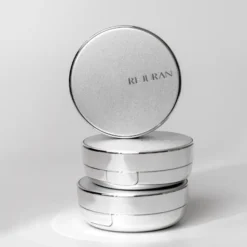 Stack of three shiny silver round cosmetic tins, with the top lid tilted and 'RI JURAN' on the lid