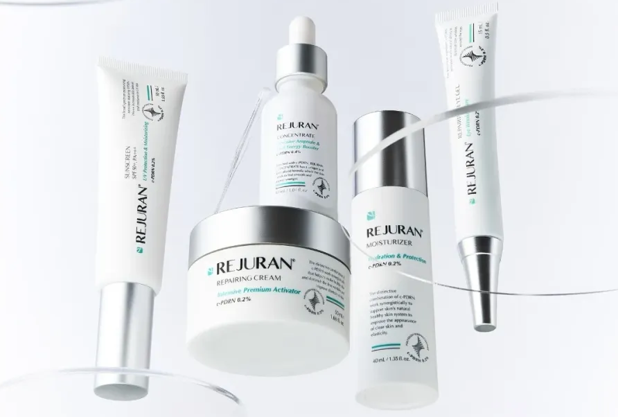 Assorted Rejuran skincare products (creams, serums, moisturizer) arranged in a line, white and silver packaging on a light background.
