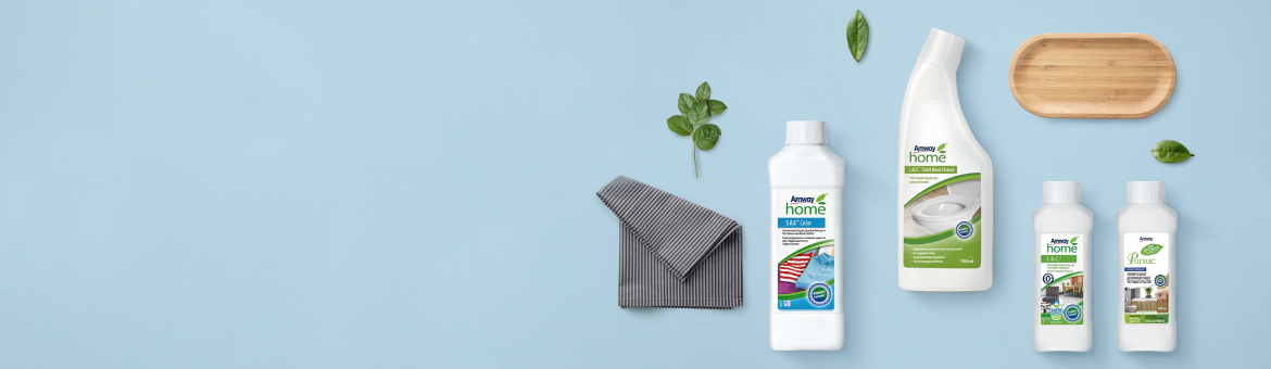 Amway Home cleaning products lined up on a light blue surface with a gray cloth and green leaves nearby.